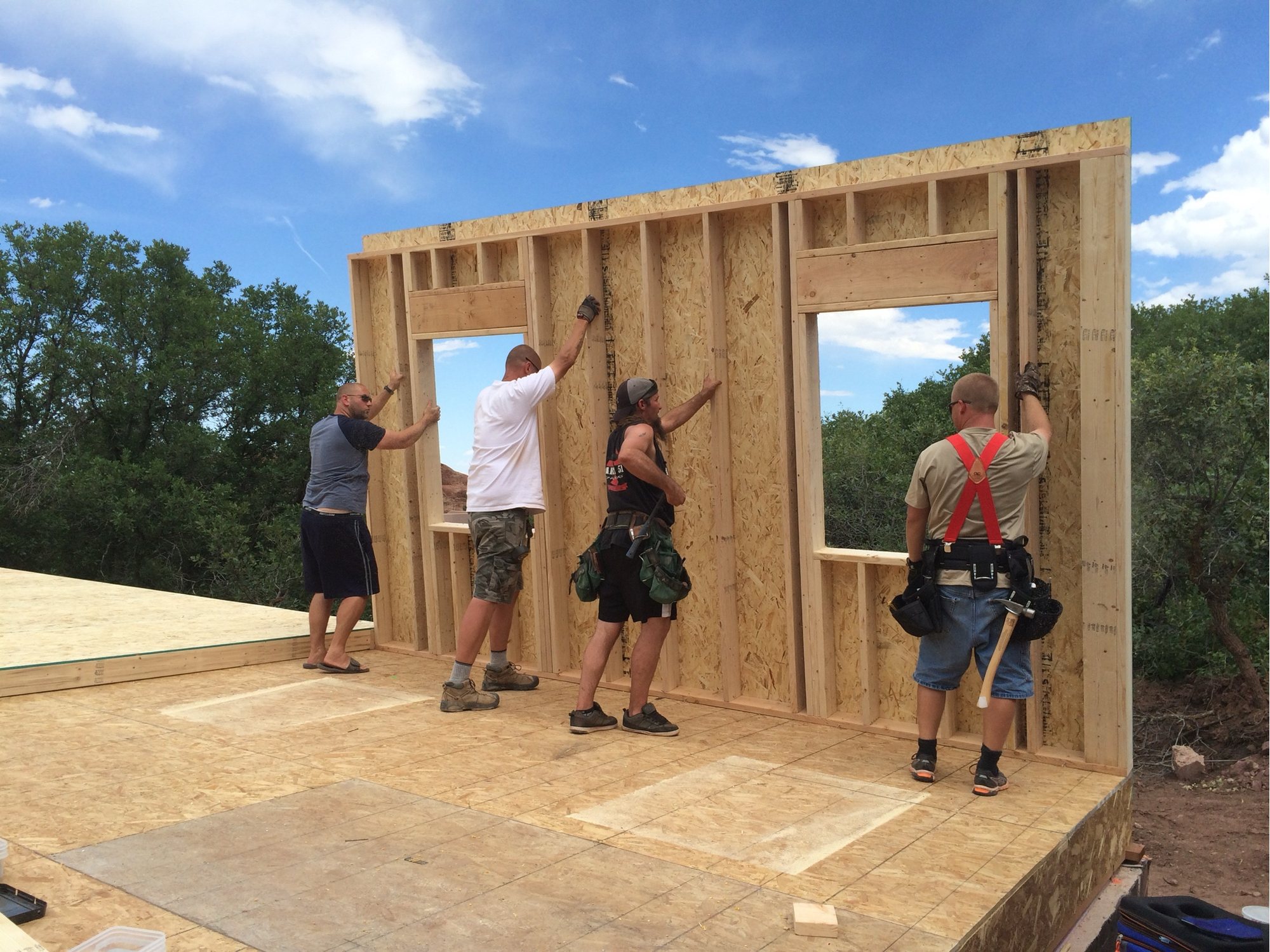 Four workers holding a newly raised framed wall upright on a building deck, red rock formations visible through the window opening